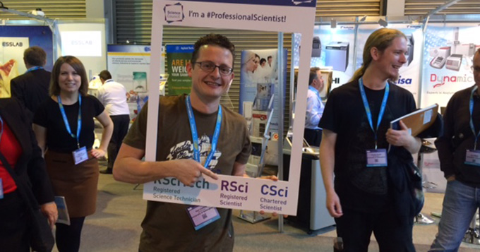 A registrant stands in a busy exhibition hall holding a professional scientist instaframe while pointing at some text on it which says 'RSci'