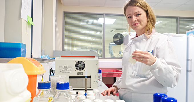 Women in white coat stands in laboratory with plastic vial