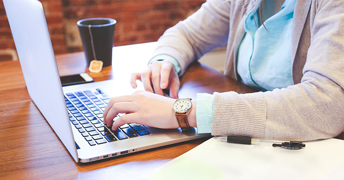 Photo of a person typing on a laptop with a notebook and pen on their desk and a mug of tea