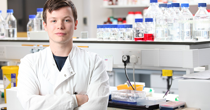 Young white man with brown hair wearing a white coat stands with arms folded and proud smile on his face in front of a laboratory bench
