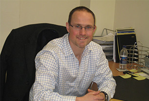 Tim, Chartered Scientist, sits with a smile at an office desk with his hands rested on the desk