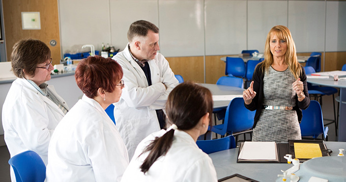 A woman who is not wearing a lab coat delivers a talk to a small group of people wearing lab coats