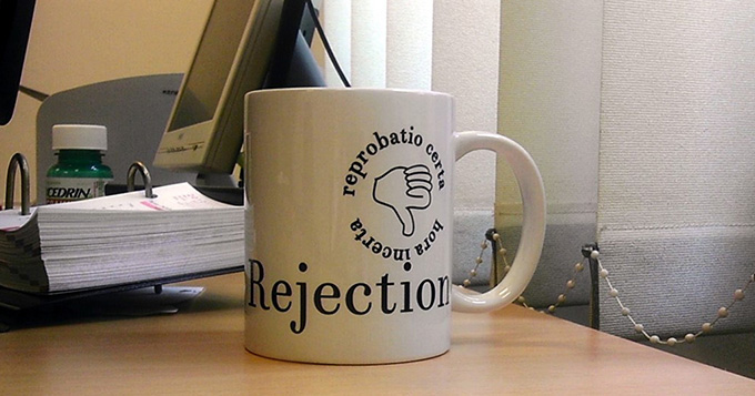 a mug on a desk - the mug has 'Rejection' written on it and a thumbs down