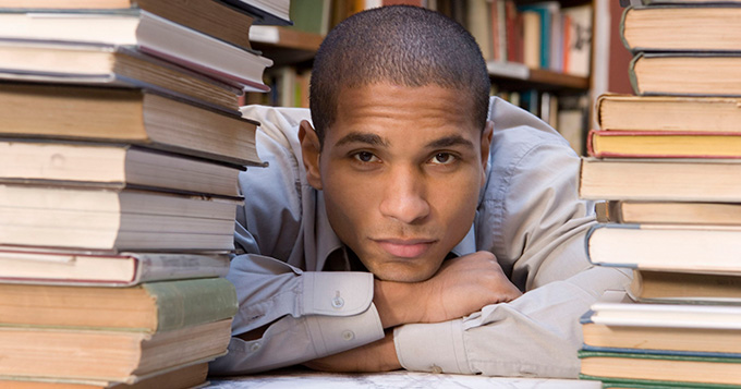 A man rests his arms and chin on a table with a stack of books piled on either side of him