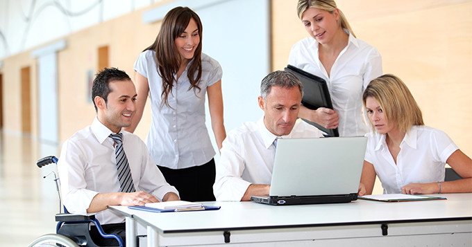 A diverse team which includes a team member sitting in a wheelchair working together around a laptop