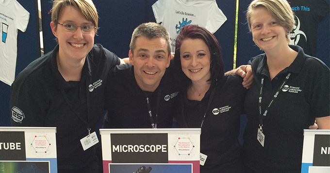 Four people wearing black SLS polo shirts standing behind a sign saying Microscope