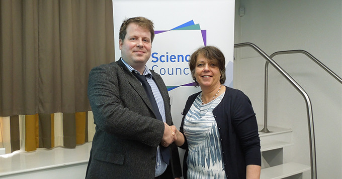 Adam Hughes, Head of Group Projects and Governance at Biochemical Society shakes hands with Belinda Phipps, Chief Executive at the Science Council in front of a Science Council logo banner