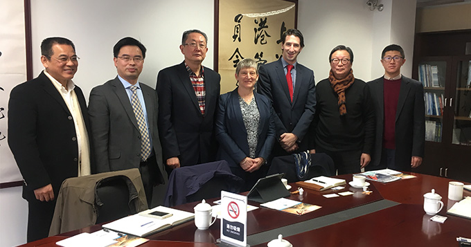 Five Chinese men, a white English woman and man all standing in a row behind a table., smiling.