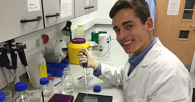 Young white man with dark hair, in a lab coat with a pipette, smiling at the camera