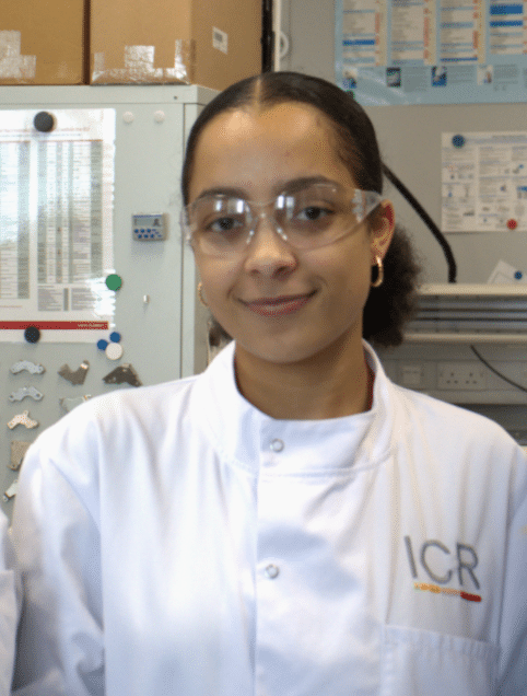 Amin smiling to camera in white lab coat with the Institute of Cancer Research logo. Amin is wearing safety glasses and has their hair tied back,