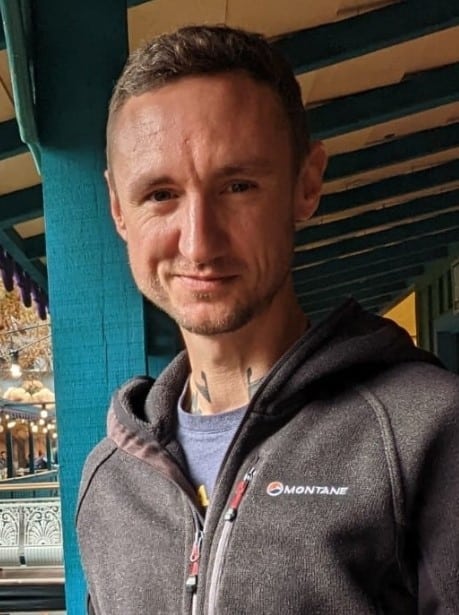 Daniel smiling to camera. They have short brown hair and are wearing a blue t-shirt with a black zipped up fleece.