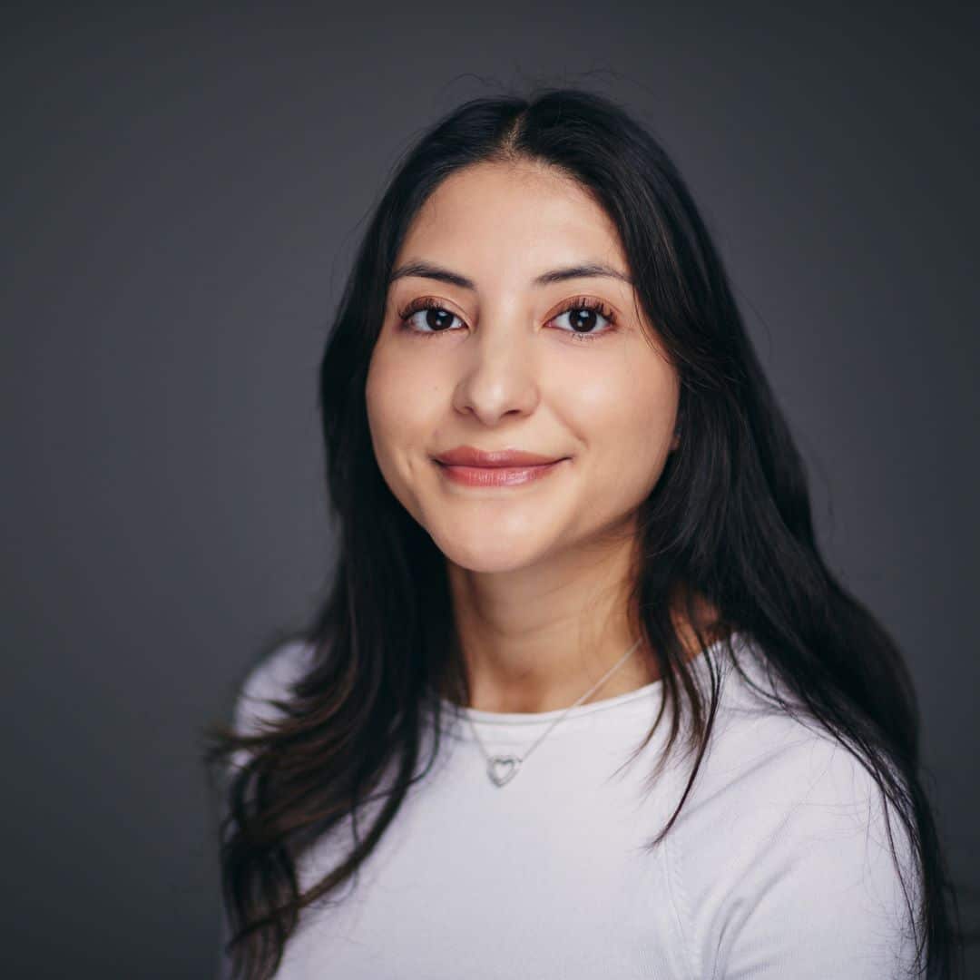 Emilia smiling to camera against a dark grey background. They have long brown hair and are are wearing a white top.