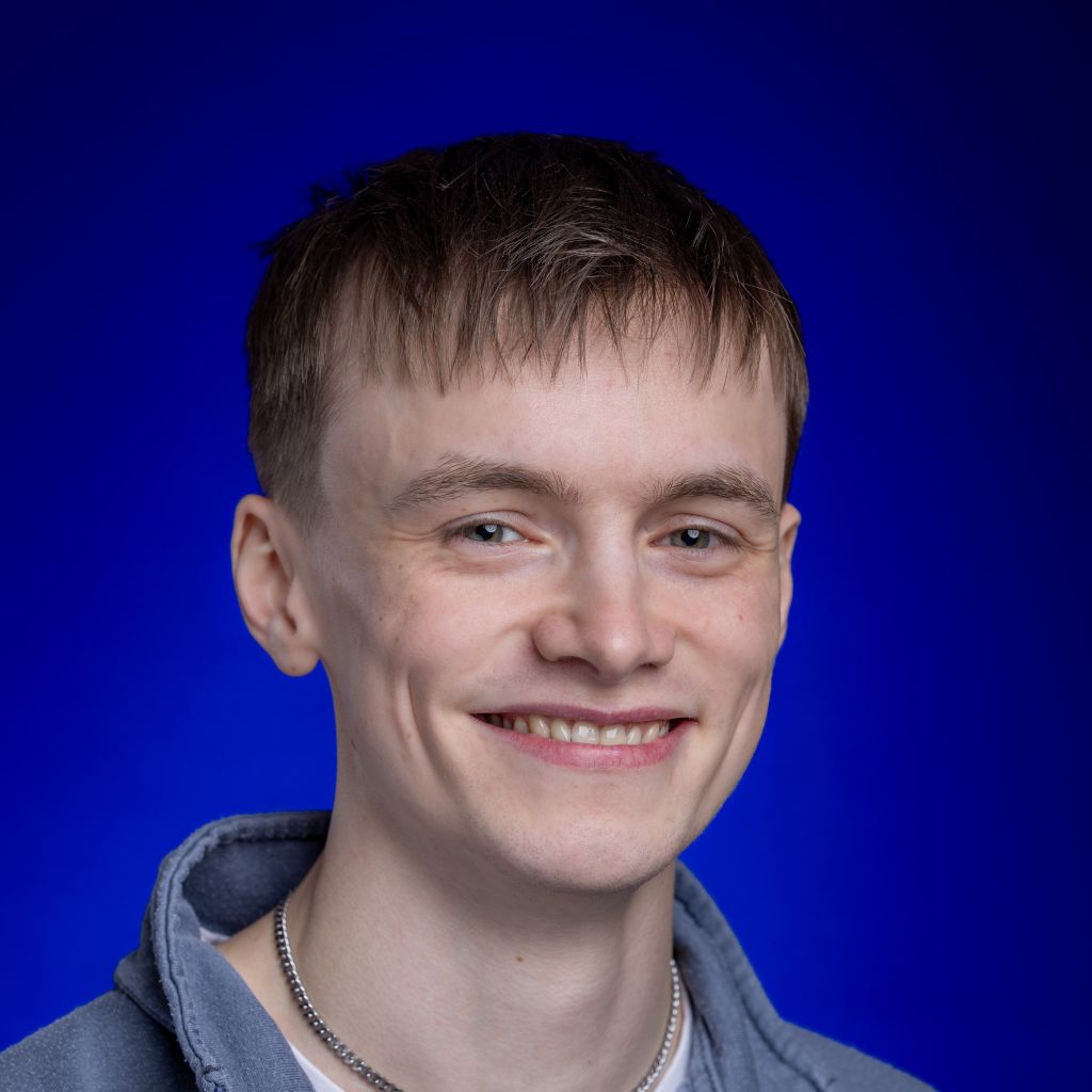 Young man with brown hair and blue eyes smiling at the camera. He wears a blue jumper with a white t-shirt and a silver chain necklace. Andrew is against an ombre royal blue background.