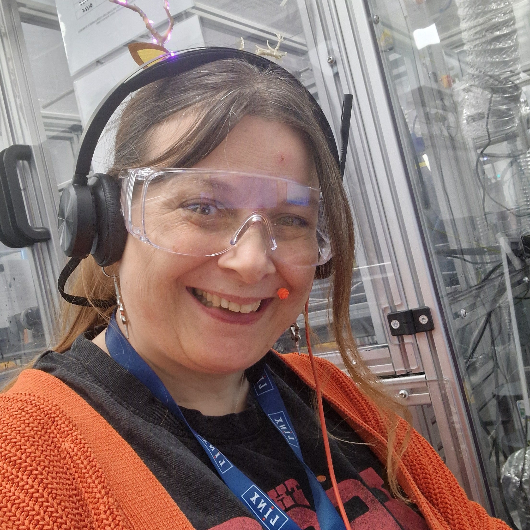 Rae is a white women with brown hair that is tied back in a pony tail. She wears a black t-shirt with a bright orange cardigan and a blue lanyard. On her head she wears black headphones, safety glasses and has a facial piercing above her lip that matches the colour of her cardigan.