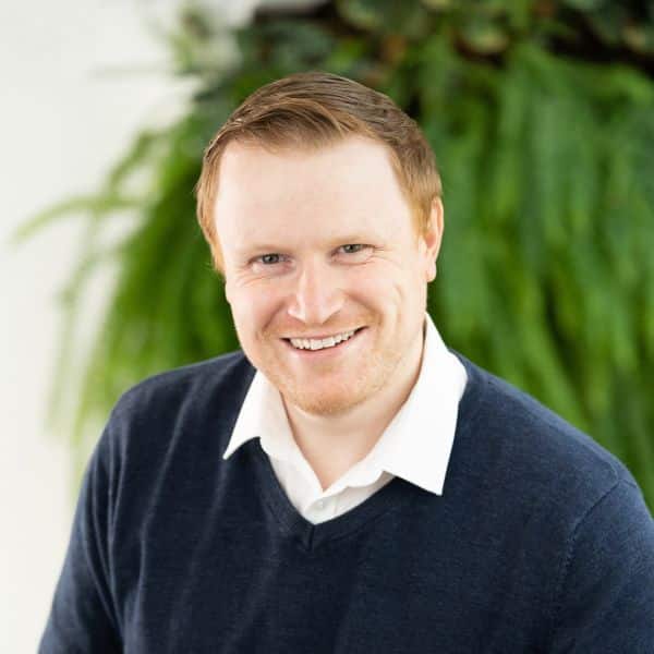 Ian has short ginger hair and wear a dark blue sweater with a white shirt. In the background is a large out of focus plant.