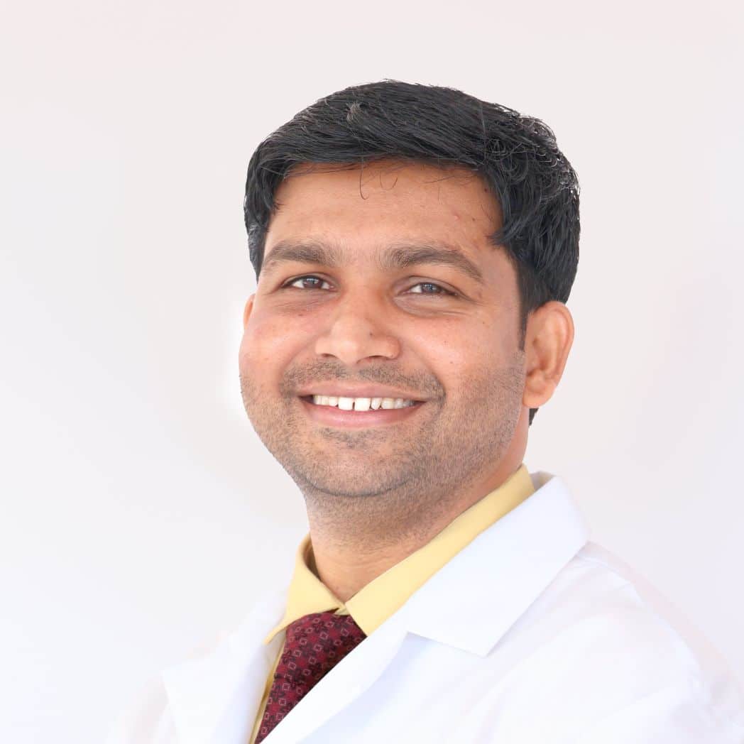 Mayank has short dark brown hair and brown eyes. He smiles to camera in a yellow shirt with red tie and white lab coat over the top.