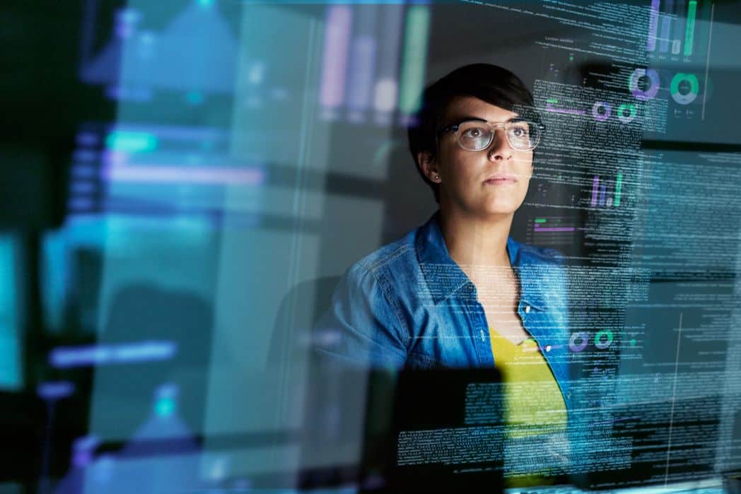 Woman with short brown hair and glasses sits behind a translucent screen that displays different types of data and graphs. She wears a yellow t-shirt and denim jacket.