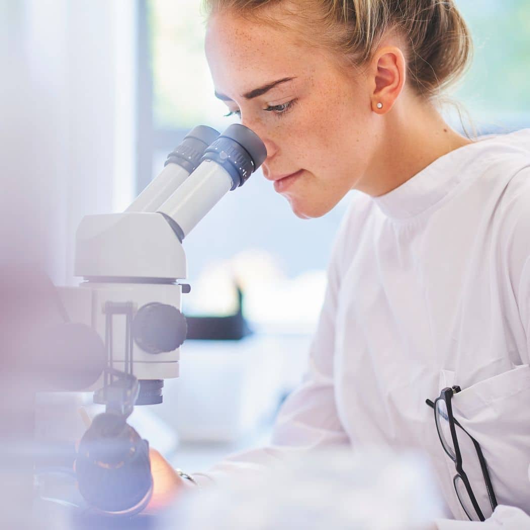 Young women looking through microscope eye pieces in a white lab coat.