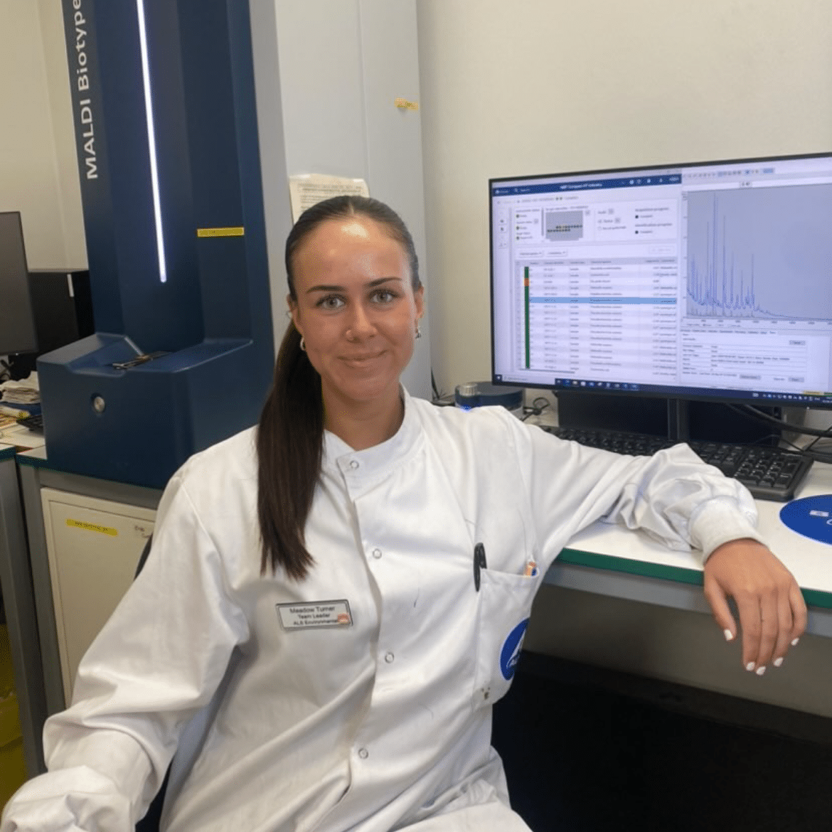 Woman in white lab coat leans back against a desk smiling to camera. A computer displaying data and lab equipment are in the backgrounhd.
