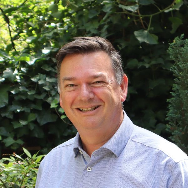 Stephen wears a light blue shirt and is smiling to the camera. In the background there is a lot of green foliage.